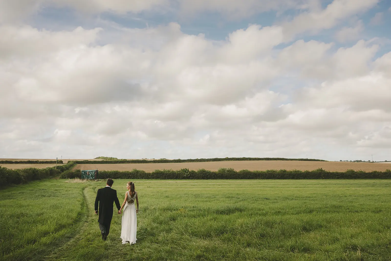 Bride and groom walking hand-in-hand through an open green Dorset field seen from behind, a vast dramatic cloudy sky stretching above the rolling farmland