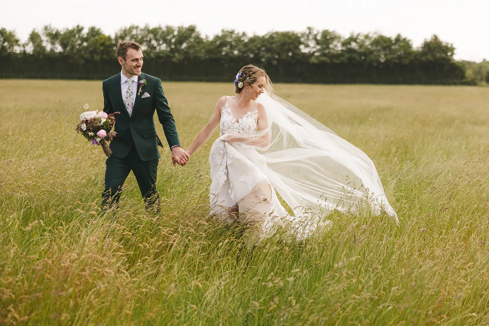 Bride and groom walking hand-in-hand through a Dorset summer meadow, bride's veil trailing in the breeze, groom in a bottle green suit with a floral tie