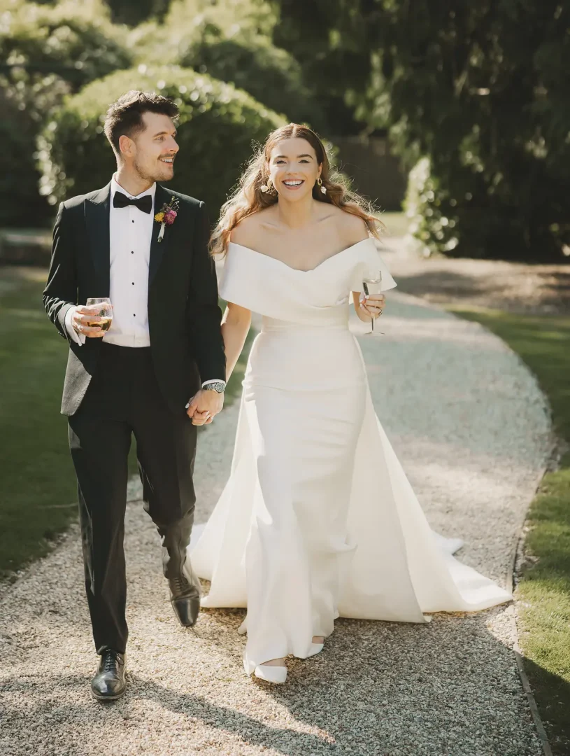 Bride in an off-the-shoulder fitted gown and groom in black tie walking laughing along a sun-dappled gravel path through the gardens at Larmer Tree, both holding champagne glasses