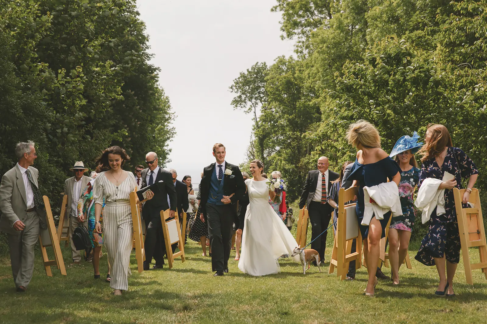 Bride and groom walking through guests carrying folding chairs along a tree-lined garden avenue after an outdoor summer wedding ceremony