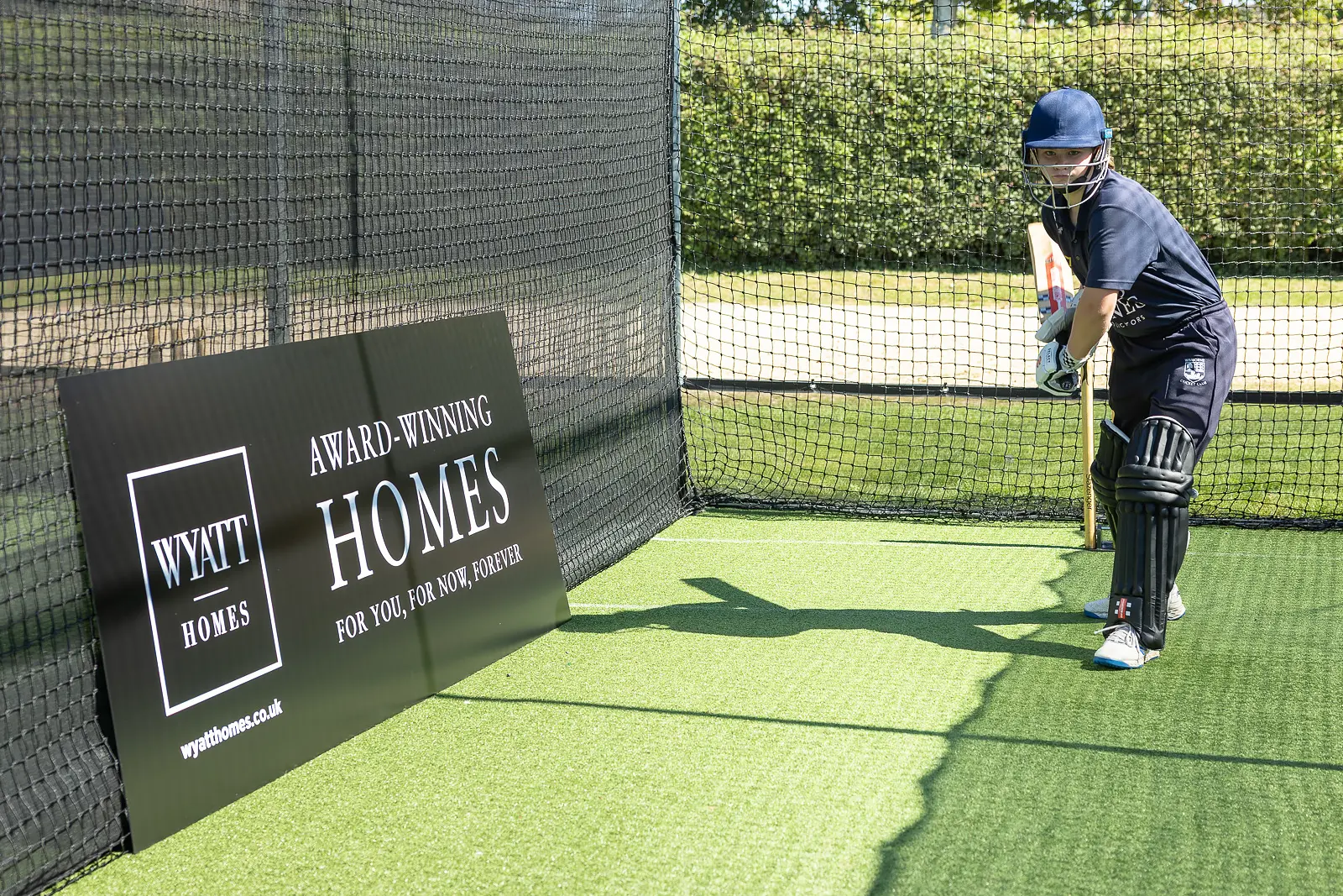 Cricketer batting in practice nets alongside a property developer sponsorship sign at a community sports event in Dorset