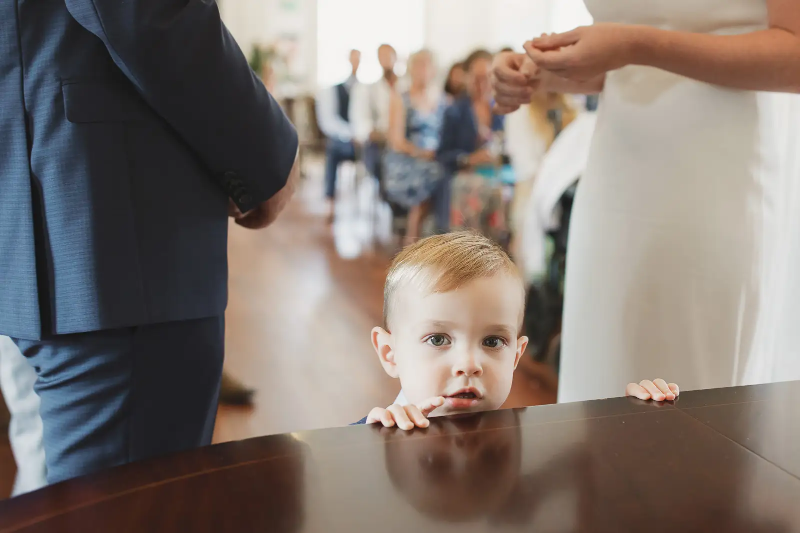 Young boy peering curiously over the signing table during wedding registry at Poole Guildhall, couple's hands visible above