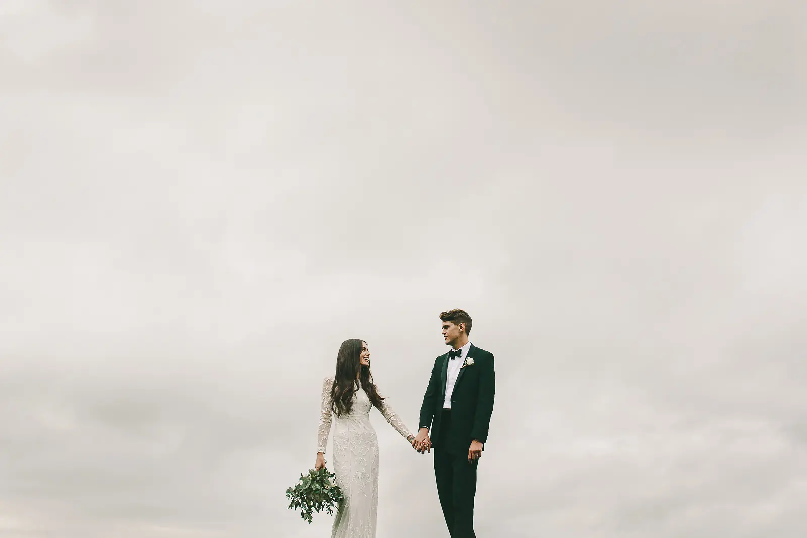 Minimalist documentary wedding portrait of bride and groom holding hands as small figures beneath a vast pale overcast sky, bride in long-sleeved lace gown