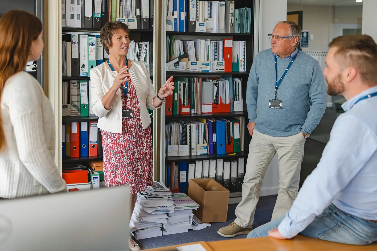 Woman leading a team discussion in a Dorset office, gesturing naturally to three listening colleagues beside shelves of files
