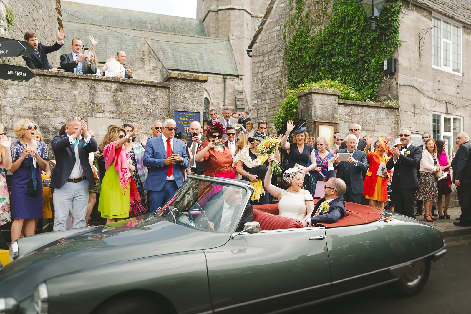 Newlyweds leaving their church wedding in Corfe Castle, Dorset, in a vintage car as guests celebrate outside