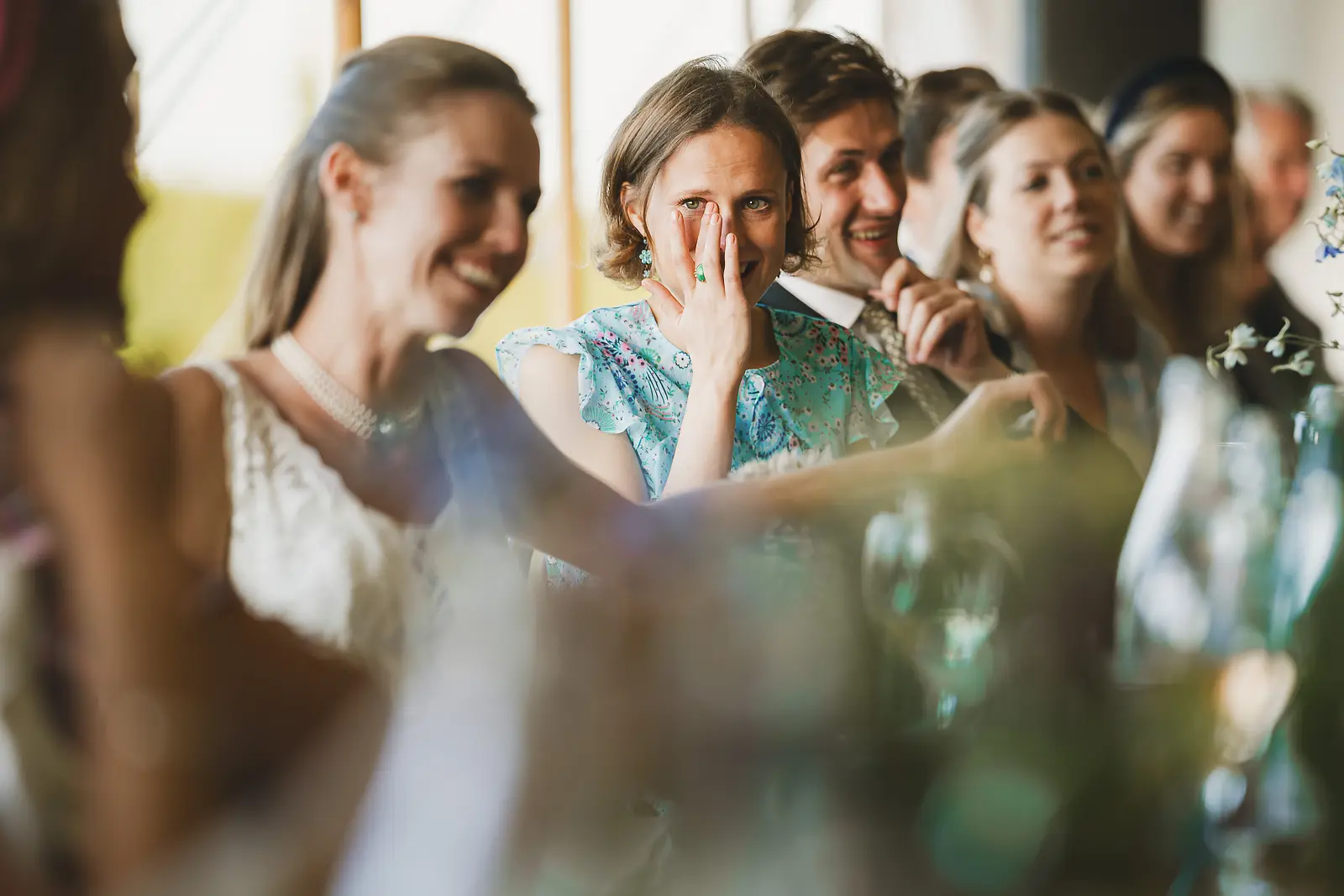 Emotional wedding guest wiping tears from her eye during the speeches at a marquee wedding reception, surrounded by smiling and laughing guests