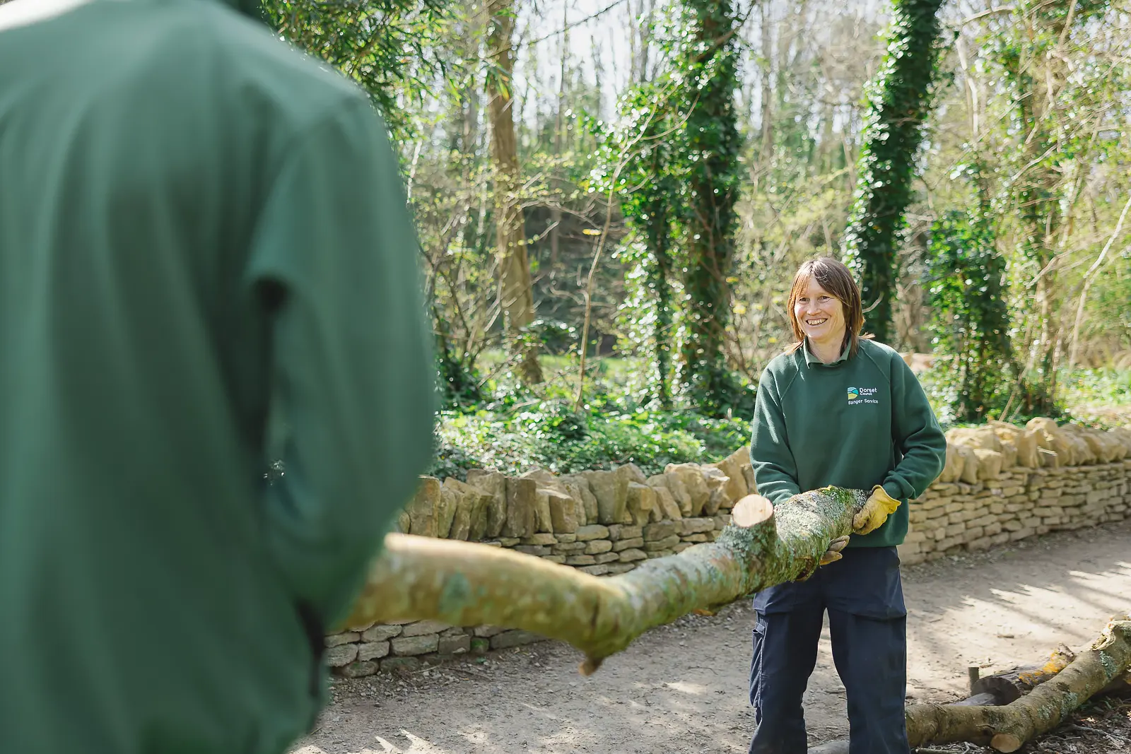 Grounds team members carrying a log together through dappled woodland at Durlston Country Park, Dorset, smiling naturally