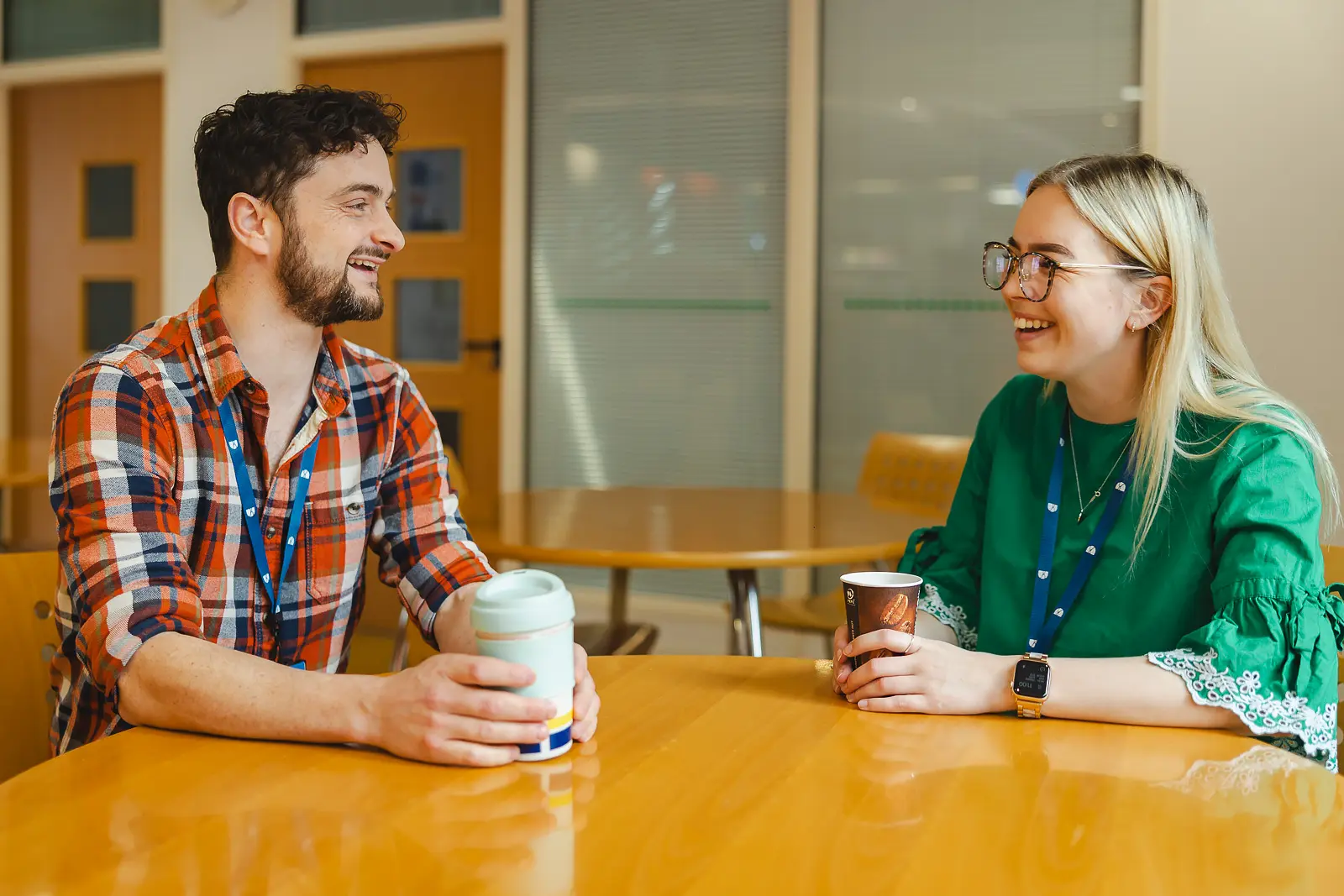 Two young colleagues laughing together over coffee in a workplace canteen in Dorset, natural and relaxed employer brand shot
