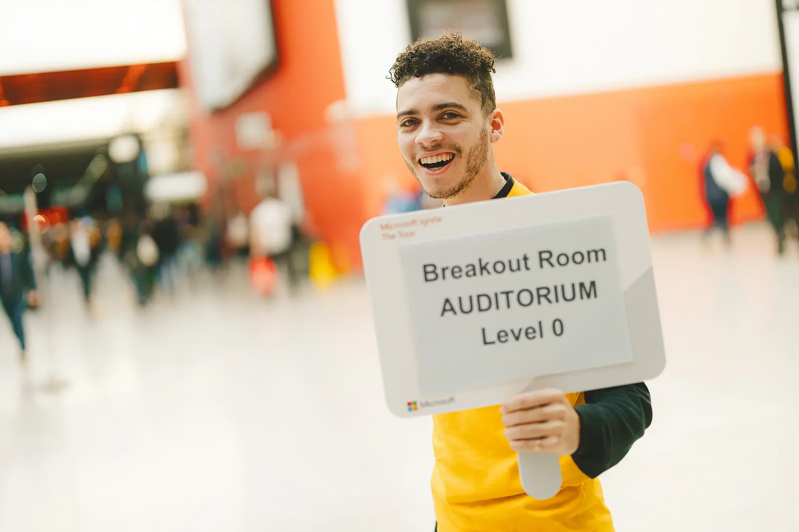 Smiling event staff member in yellow uniform holding a breakout room auditorium direction sign in ExCeL London venue