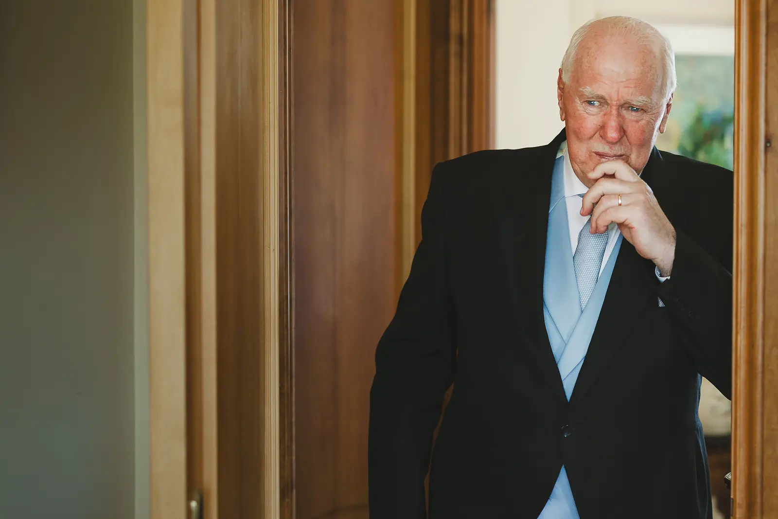 Documentary portrait of an older gentleman in morning suit with pale blue cravat standing in a doorway, hand to chin in quiet contemplation before the wedding ceremony