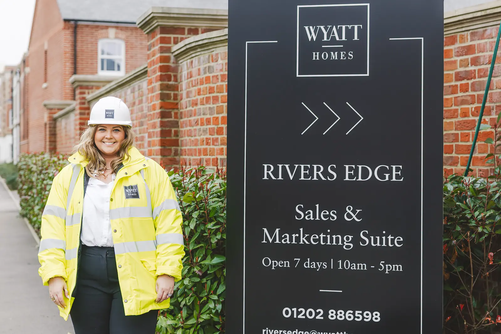 Smiling female staff member in hi-vis jacket and hard hat standing beside a property development sales and marketing suite entrance sign in Dorset