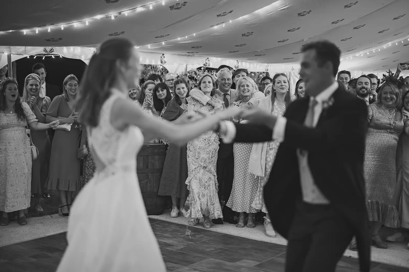 Black and white first dance with joyful wedding guests looking on and cheering from the edge of the dance floor, fairy lights strung across the marquee ceiling