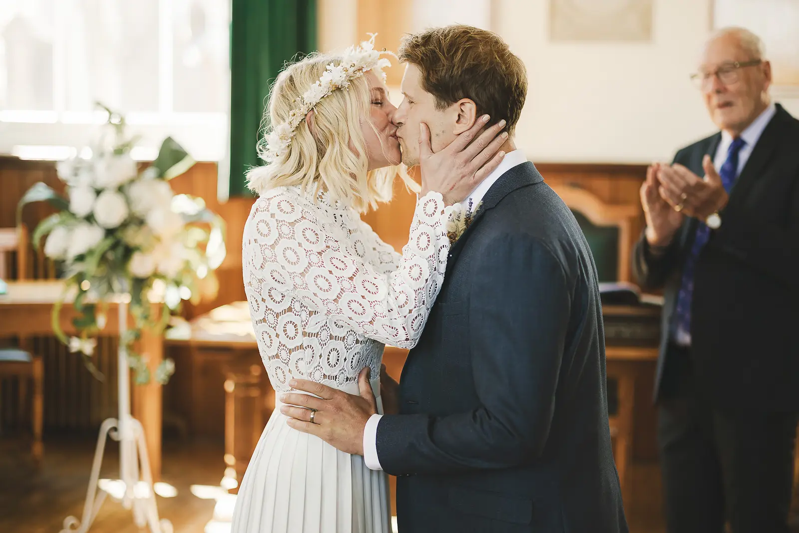Bride in a lace top and dried flower crown cradling her groom's face for their first kiss at a registry office wedding ceremony, registrar clapping with joy behind them