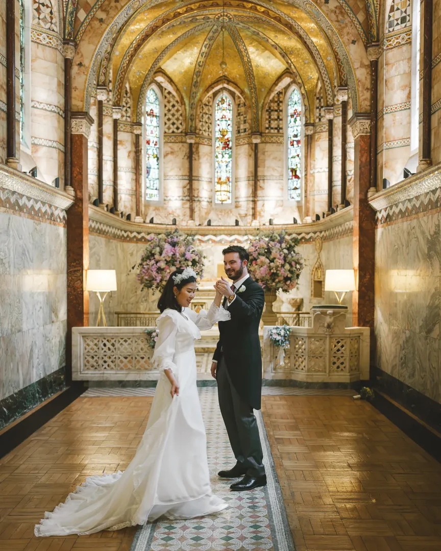 Bride and groom exchanging rings inside the ornate Fitz Rovio Chapel in London, with gilded Byzantine mosaic apse, marble columns and a pink floral arch behind them