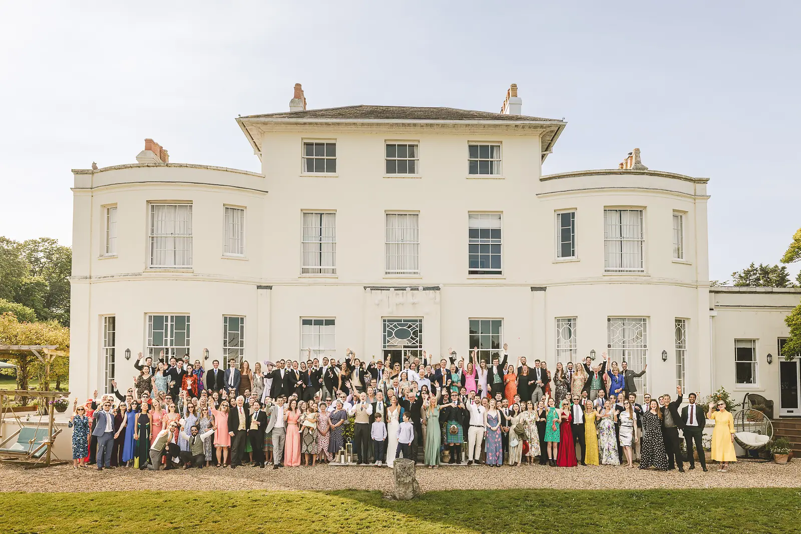 Full wedding group photograph with all guests gathered on the lawn in front of the grand white Regency facade of Studland Bay House in Dorset