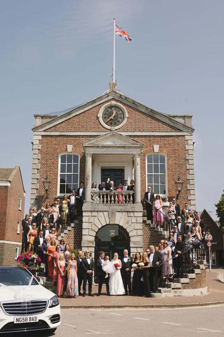 Full wedding party and guests gathered on the grand exterior steps of Poole Guildhall Registry Office, Union Jack flying above the clock tower on a sunny summer day