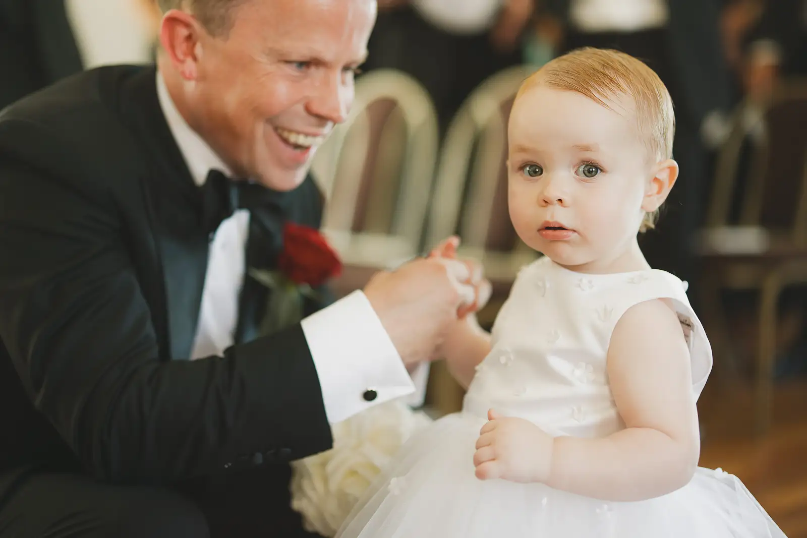 Groom in black tie laughing as he holds hands with a wide-eyed baby in a white dress during Poole Guildhall registry office wedding