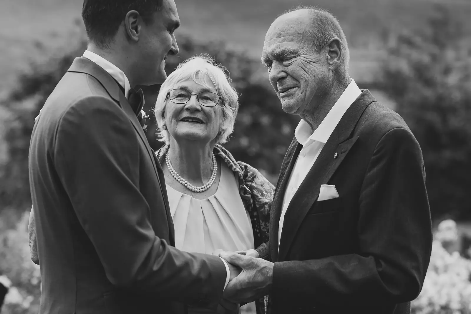 Black and white candid of a groom holding hands with his elderly parents at a wedding, his mother smiling warmly in a pearl necklace, his father moved with emotion