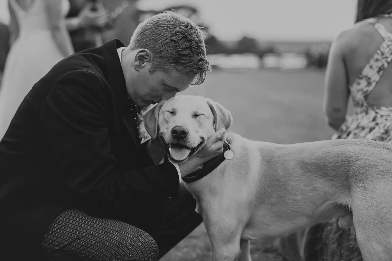 Candid black and white moment of a groom crouching to nuzzle a contented yellow Labrador at an outdoor wedding reception on a summer evening