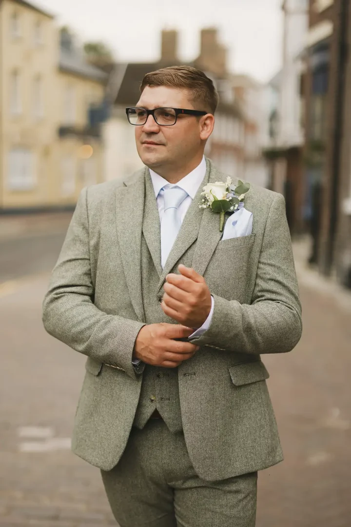 Dapper groom in sage green herringbone three-piece suit and white rose buttonhole on Poole Old Town street before wedding