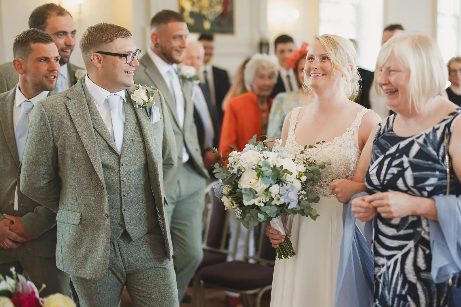 Groom turning to smile as his bride arrives at the altar inside Poole Guildhall, guests standing and watching behind