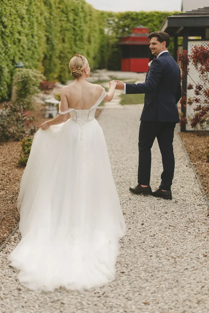 Groom spinning his bride on a gravel garden path lined with tall clipped hedges, bride in an off-shoulder tulle ballgown revealing the button-back corset detail