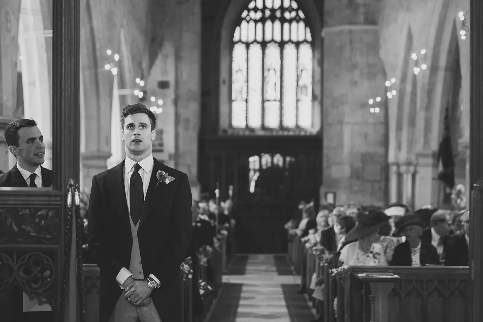 Black and white photograph of a groom in morning dress standing at the church altar in anticipation as guests fill the pews behind him