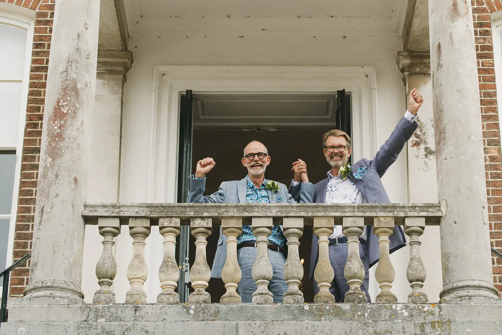 Two grooms punching the air in celebration on the balcony of Poole Guildhall after their registry office wedding