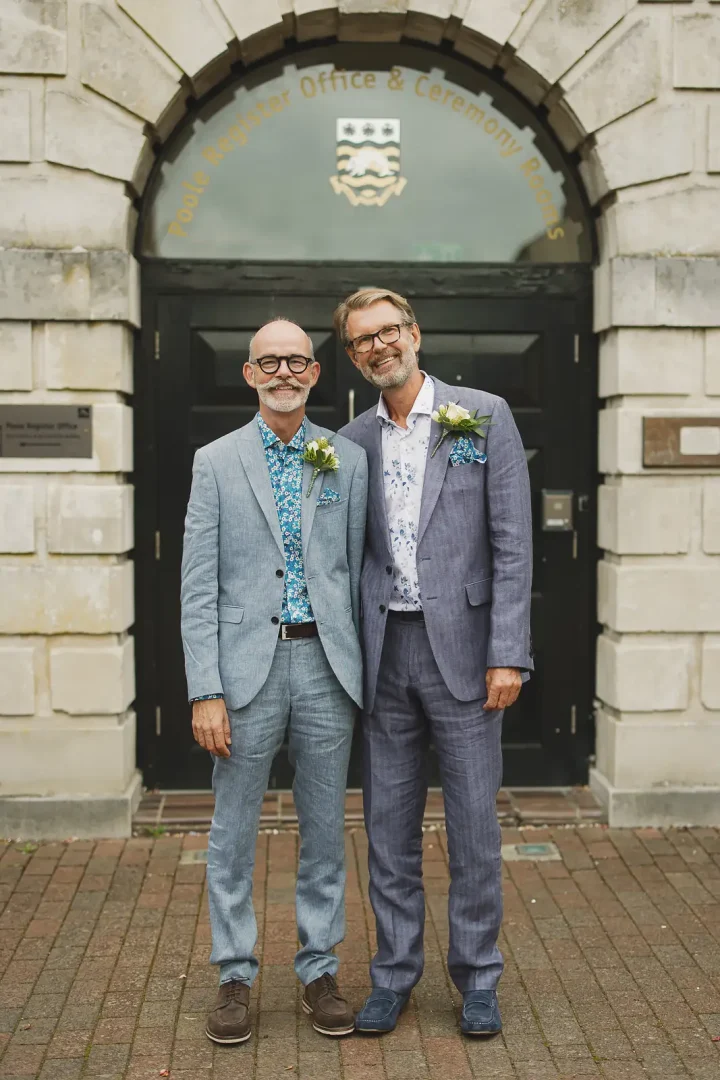 Two grooms posing together outside Poole Guildhall Registry Office entrance in matching light grey suits with flower buttonholes