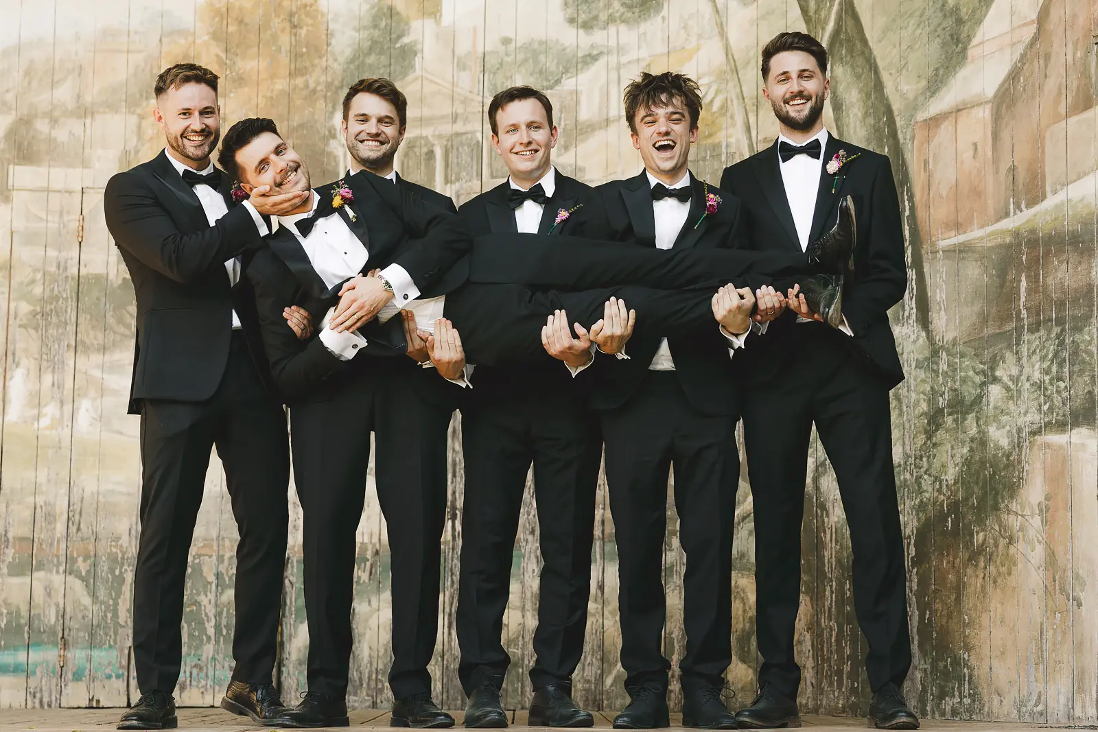 Groomsmen in black tie lifting the groom horizontally while he laughs with delight, in front of the painted scenic backdrop at Larmer Tree Gardens
