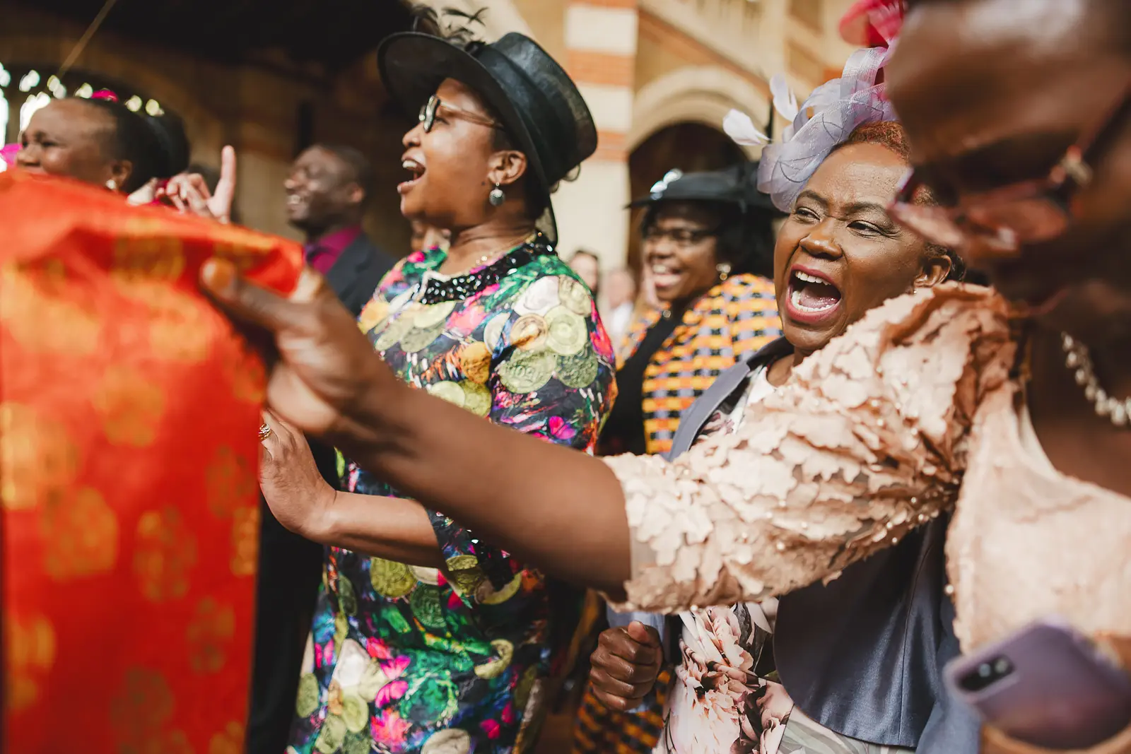 Wedding guests singing and celebrating with abandon in vibrant colourful outfits and hats, including kente-patterned fabric, outside a Bournemouth church wedding