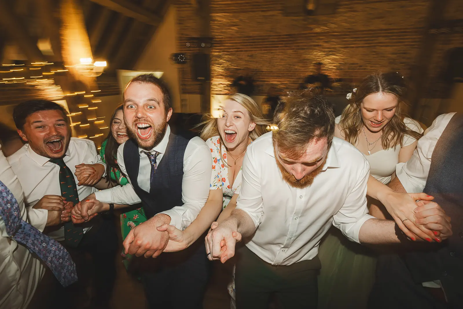 Wedding guests screaming with laughter as they form a conga chain on the dancefloor of a warmly lit Dorset barn wedding reception