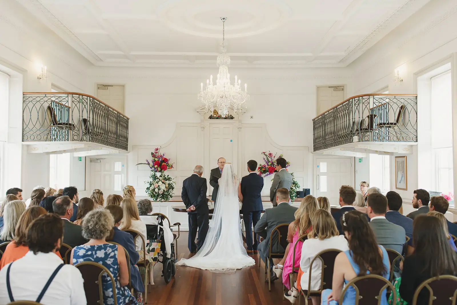 Wide view of wedding ceremony inside Poole Guildhall's elegant Georgian room with crystal chandelier and ornate balconies
