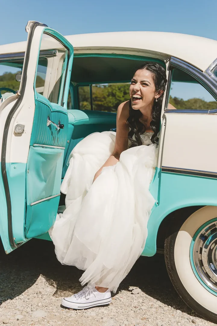 Laughing bride in a full tulle wedding dress and Converse trainers stepping out of a turquoise classic 1950s American car on a sunny wedding day