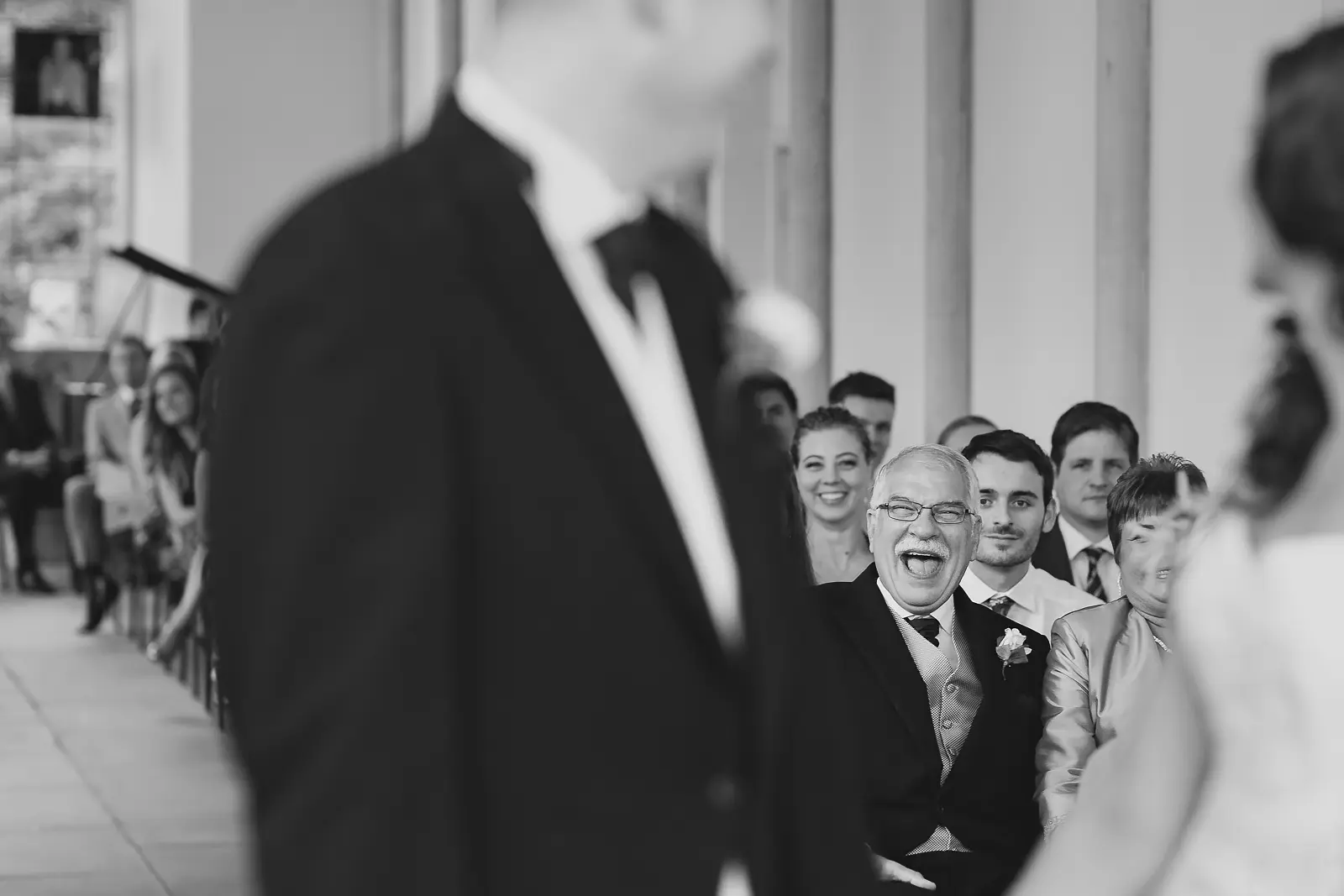 Candid black and white photograph of a laughing wedding guest in the crowd during a ceremony at a grand country house with colonnaded architecture