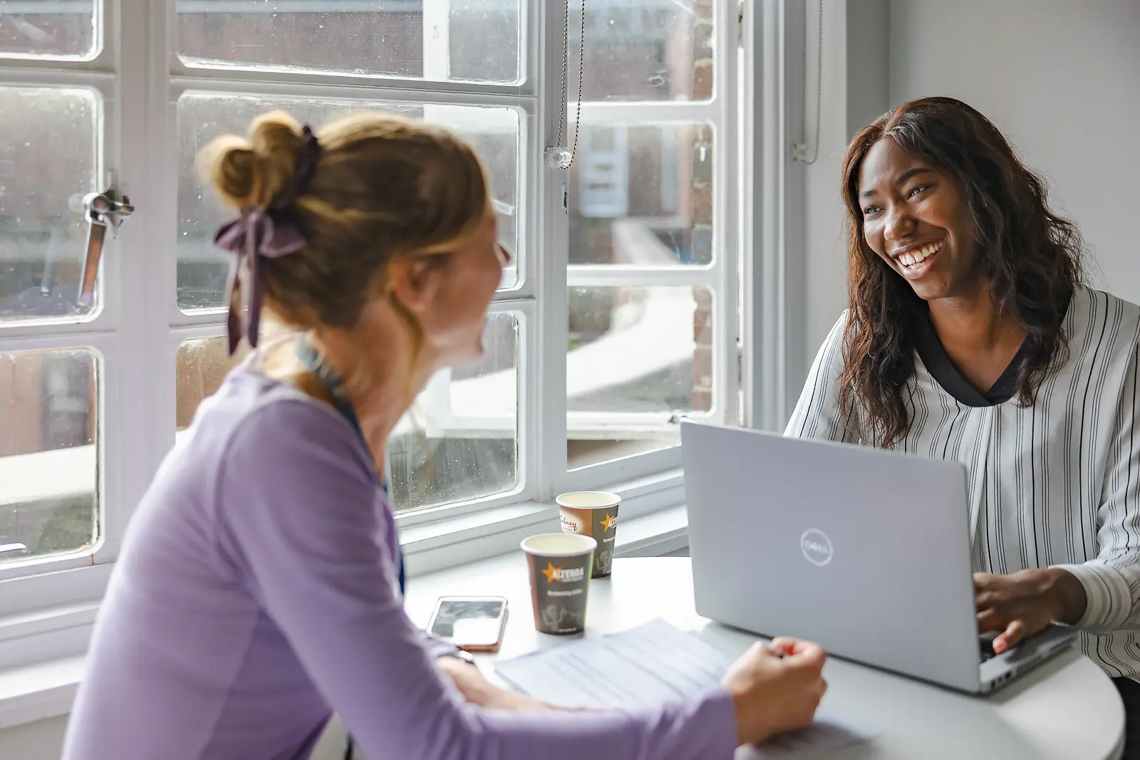 Two colleagues laughing together over coffee and a laptop at a sunny window in a Dorset office, natural and candid