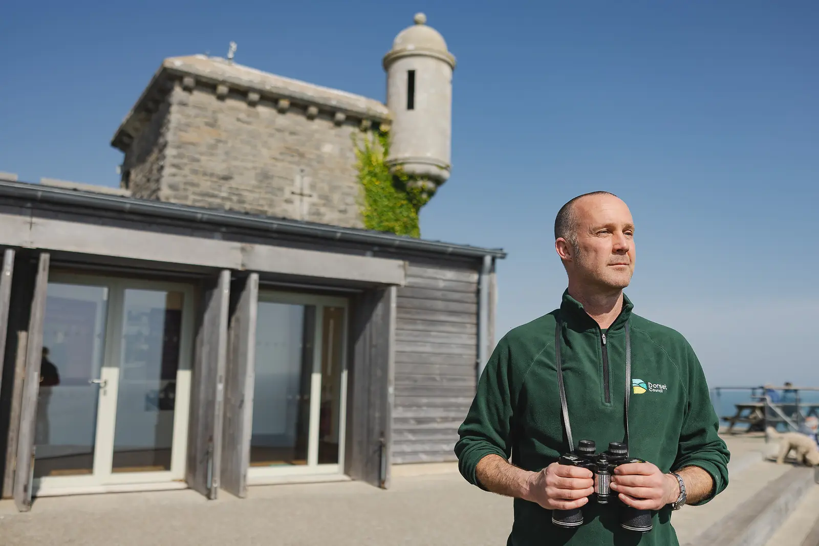 Grounds team member holding binoculars outside Durlston Castle in Dorset, on-location lifestyle portrait against a clear blue sky