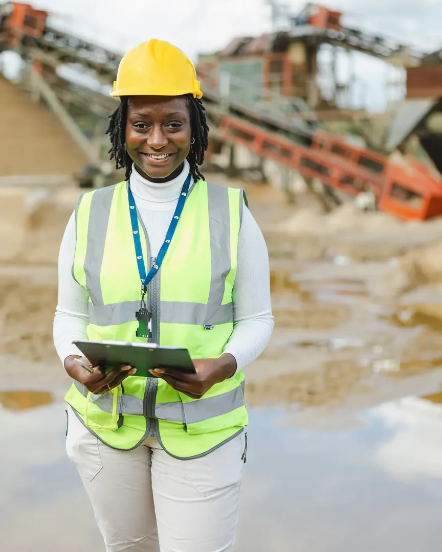 Smiling monitoring officer in hi-vis vest and hard hat holding a clipboard at Woodsford Quarry in Dorset, quarry machinery behind