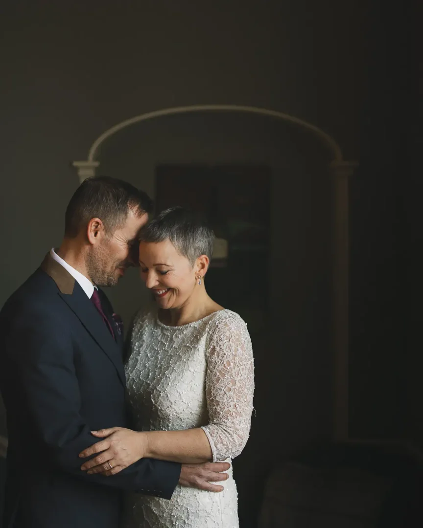 Intimate portrait of bride and groom inside Poole Guildhall, dramatic window light with arched doorway framing them in shadows