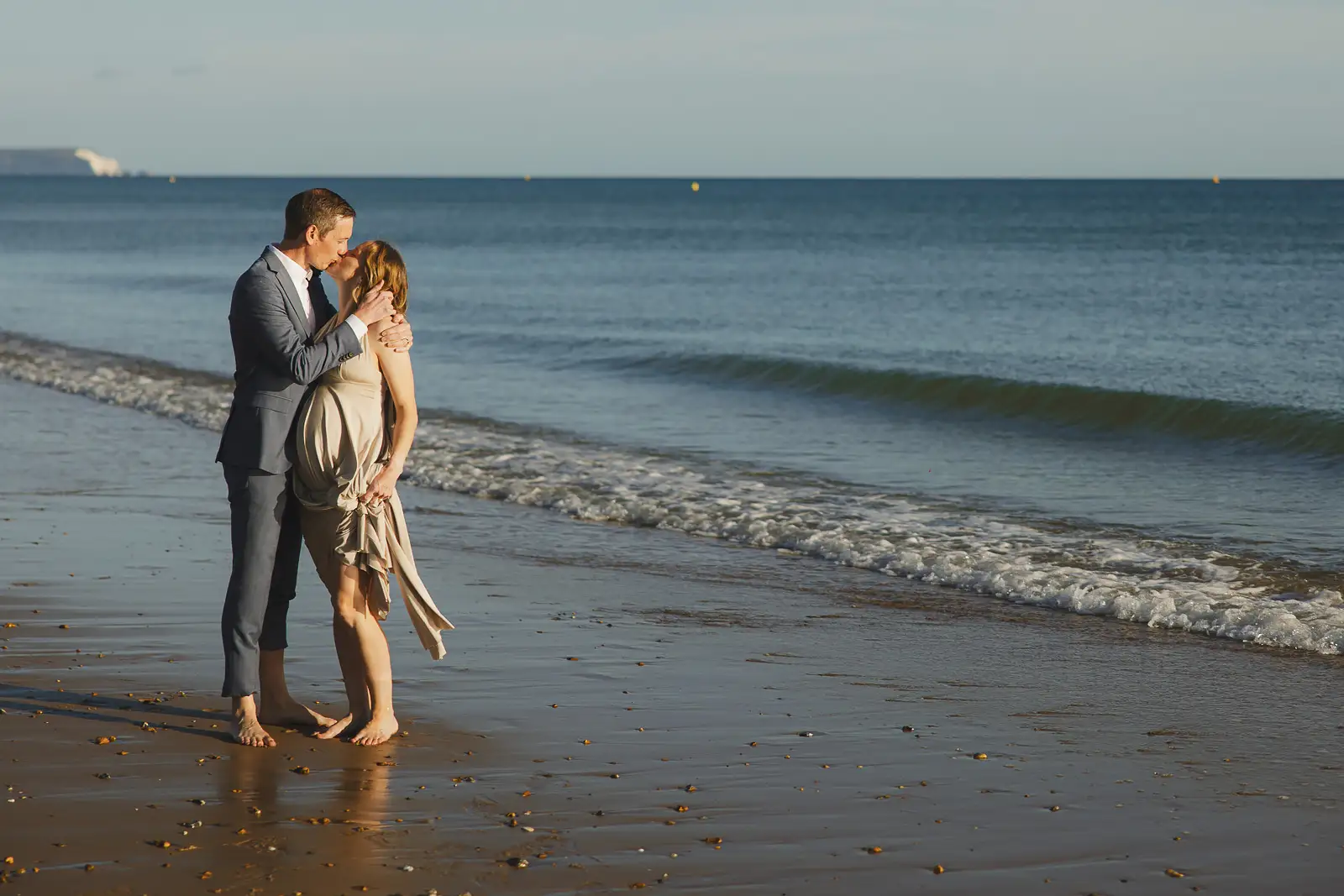 Newlyweds kissing barefoot at the water's edge on Bournemouth beach in warm golden light, waves rolling in behind