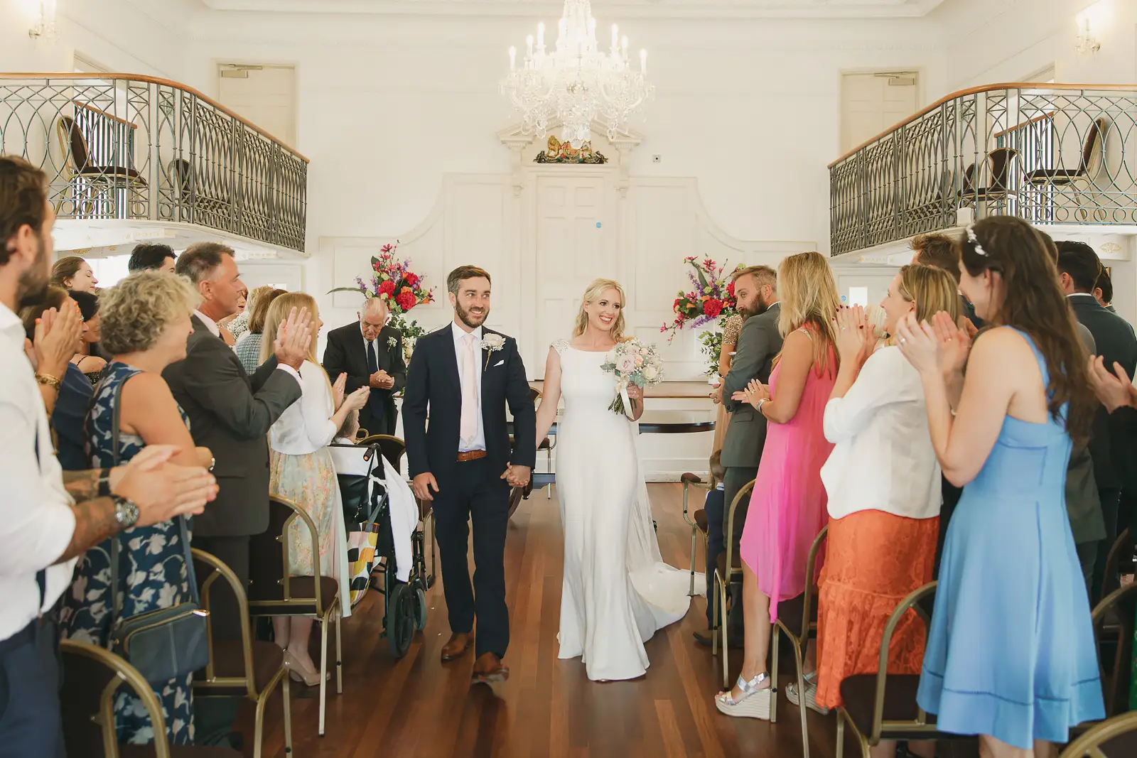 Newlyweds walking back down the aisle after wedding ceremony at Poole Guildhall as guests applaud, chandelier and balconies above