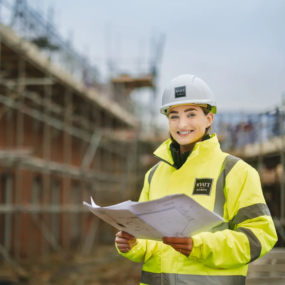 Young female staff member in hi-vis jacket and hard hat holding building plans and smiling at camera on a residential construction site