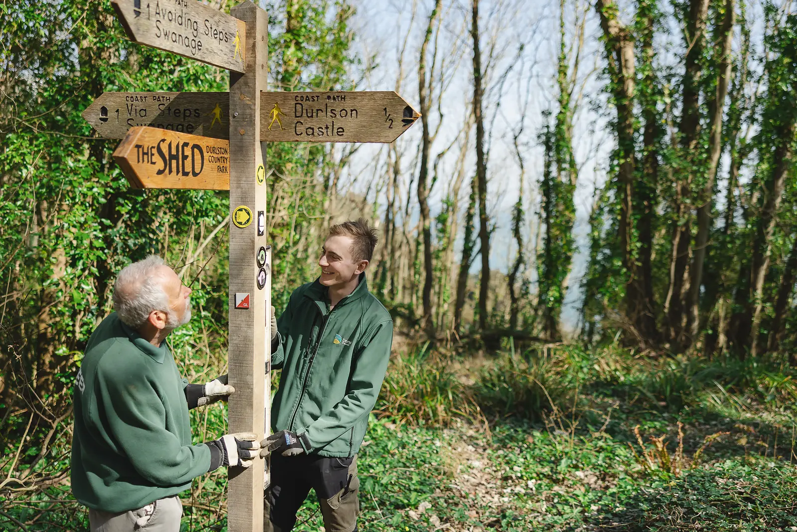 Two grounds team members in green uniform beside a coastal path signpost at Durlston Country Park in Dorset, natural and candid