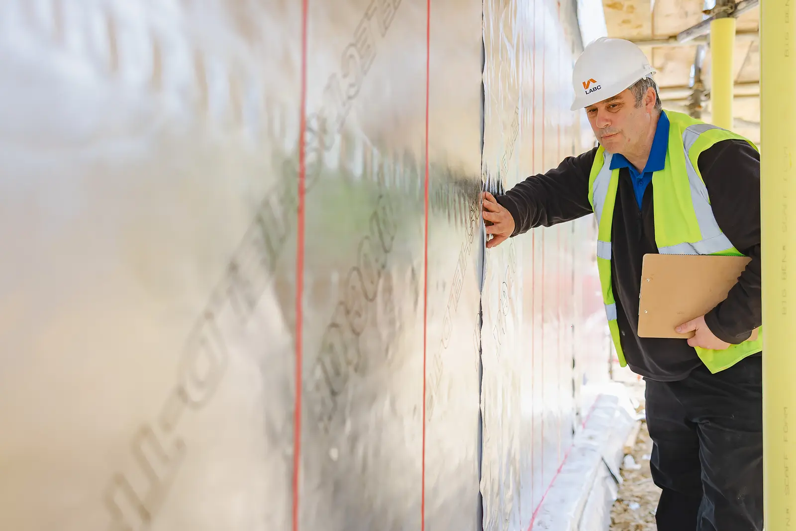 Building inspector in white hard hat and hi-vis vest examining a construction wall with clipboard at a Poundbury development site