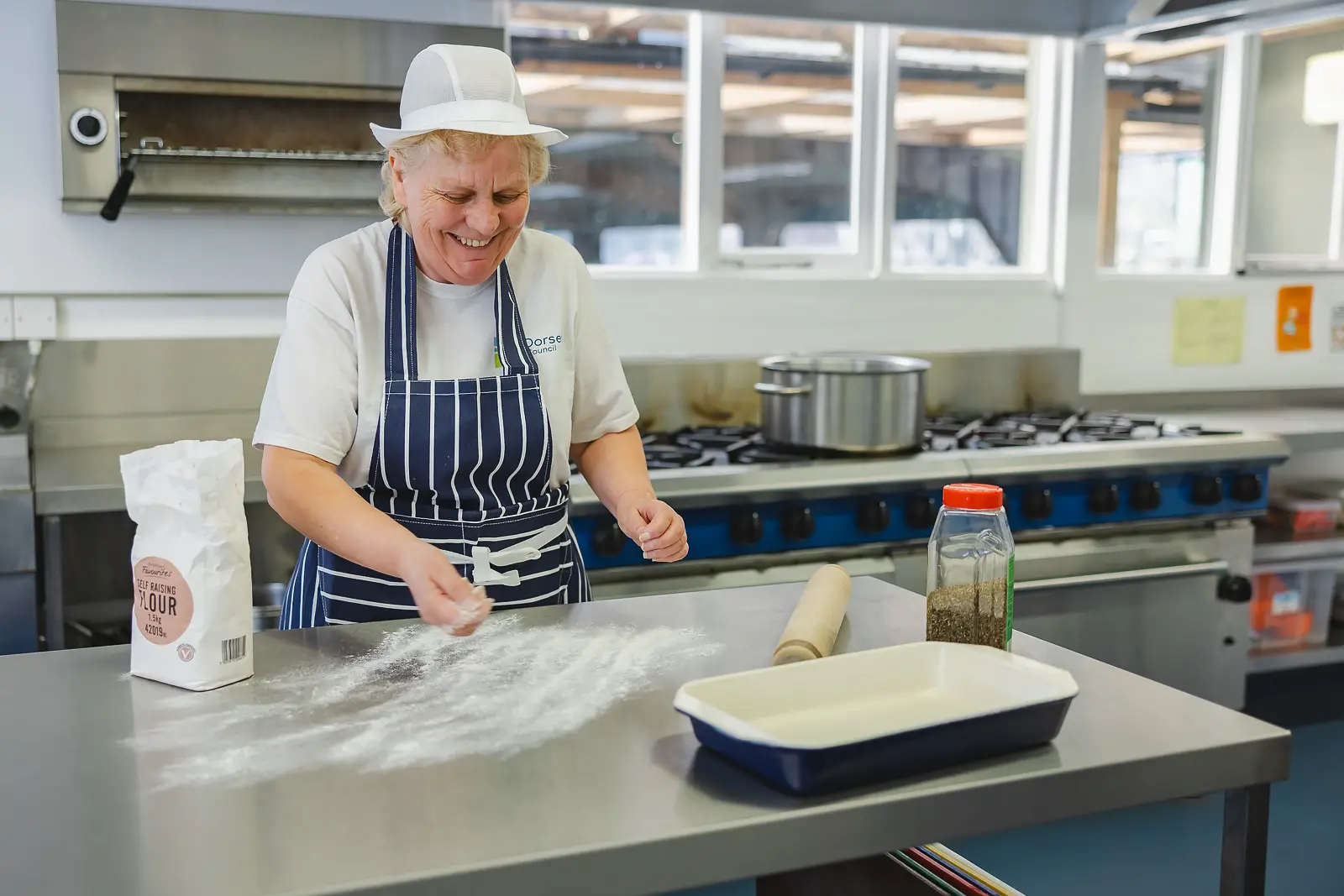 Laughing catering worker in chef's hat and striped apron sprinkling flour on a stainless steel worktop in a Dorset commercial kitchen