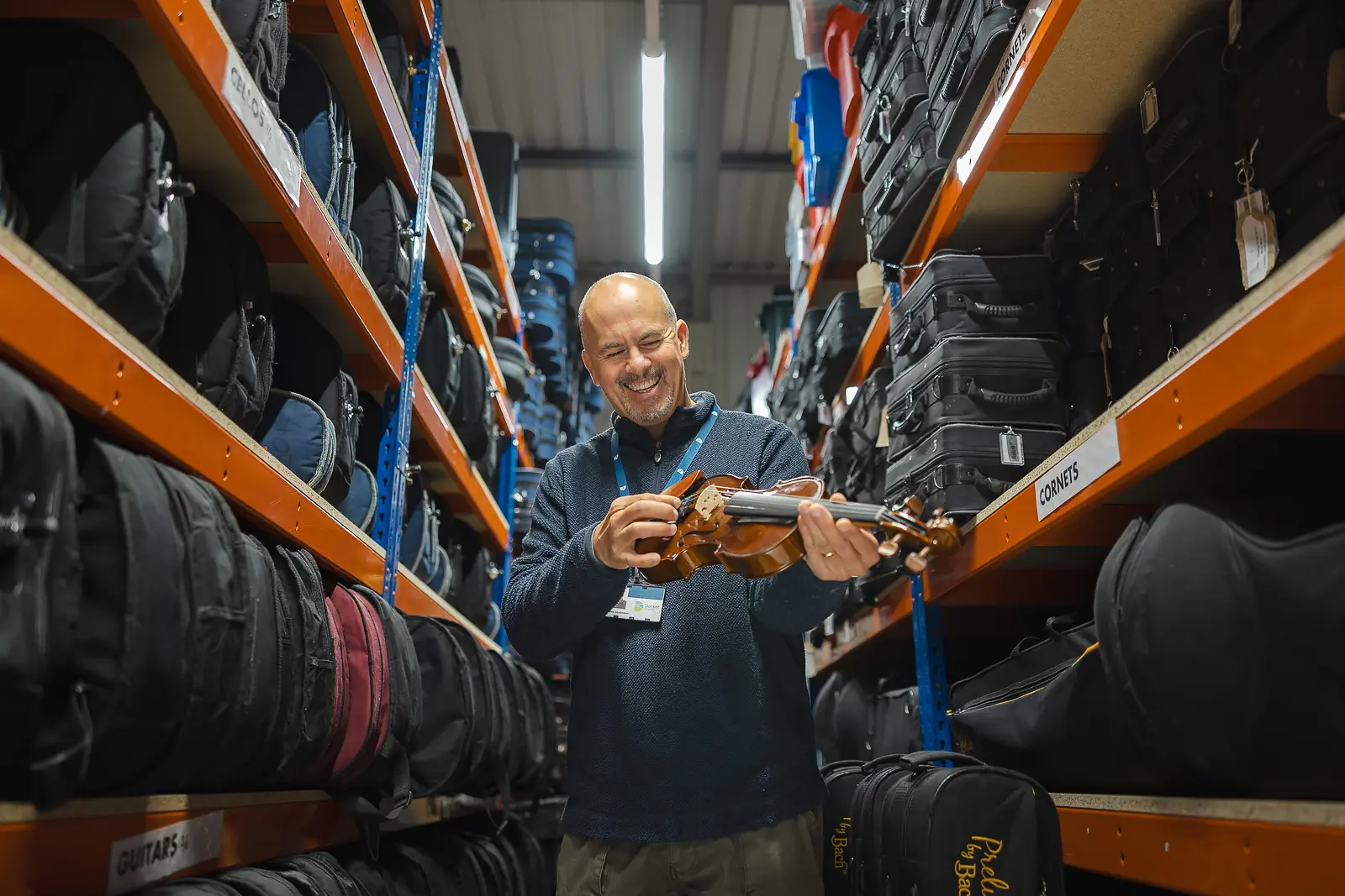 Instrument technician smiling and holding a violin in a music services warehouse stocked with instrument cases in Blandford, Dorset