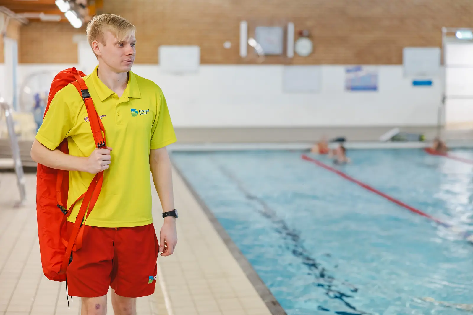 Young lifeguard in yellow uniform carrying rescue equipment beside an indoor swimming pool in Dorset, natural on-location shot