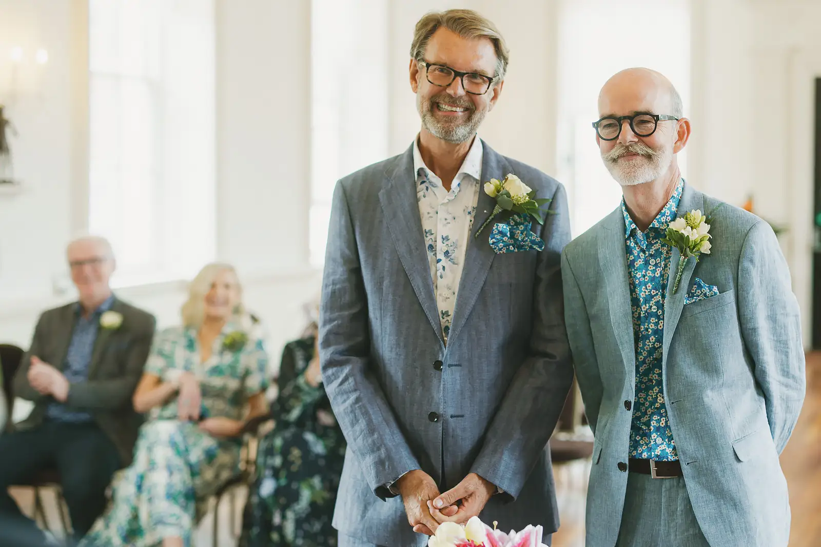 Two grooms smiling together at the start of their wedding ceremony inside Poole Guildhall, guests seated behind