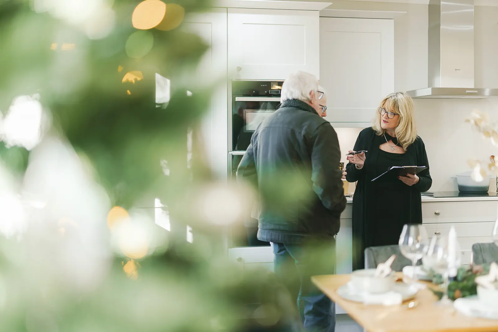 Sales advisor with clipboard in discussion with a visitor in a show home kitchen framed through a lit Christmas tree with bokeh lights in foreground