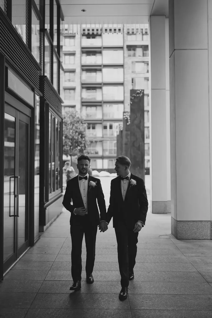 Black and white portrait of two grooms in black tie tuxedos holding hands and walking along a city pavement with a modern glass office building behind them